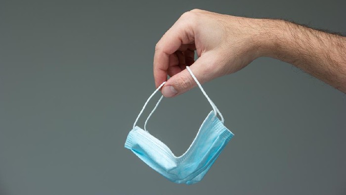 Caucasian male hand dangling a blue textile medical mask close up front view isolated on gray.