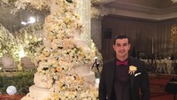 Pose mengenakan jas formal, Ali berdiri tepat di samping cake pengantin mewah dengan banyak hiasan bunga berwarna putih. Eits tapi ini bukan cake pernikahan ia.  Foto: instagram @alisyakieb