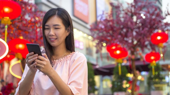 online imlek Asian woman standing at city steret decorated with lantern, sending greeting wishes with smart phone to the family and love one during Chinese new year