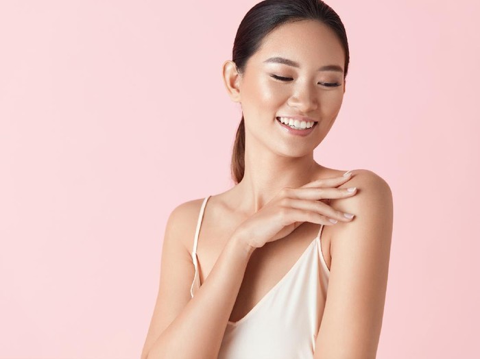 Beauty. Asian Woman Portrait. Ethnic Model With Smooth Skin Touches Her Shoulder And Smiling Against Pink Background. Confident Female Enjoying Her Perfect Body After Cosmetology Treatment.