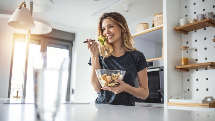 ilustrasi diet Photo of young woman enjoying a delicious salad while standing in her kitchen at home during the day.