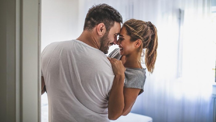 Ilustrasi Posisi Seks Handsome man in white T-shirt carrying young attractive woman while spending free time at home. Loving young hipster couple relaxing at home and kissing. View from another room through the doorway