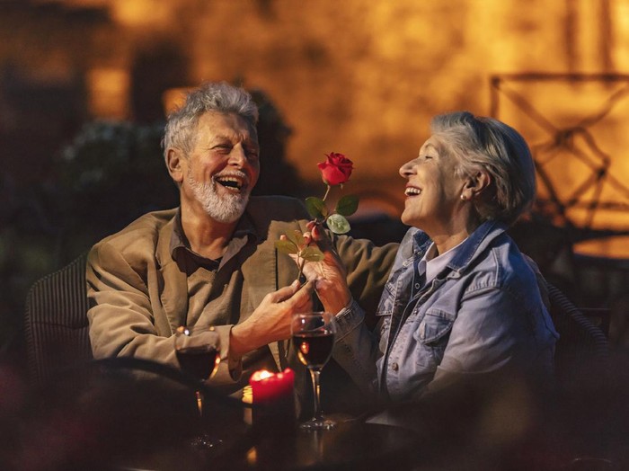 Lovely old lady admiring the red flower that her husband gave her. Couple having glass of red wine at night in roof restaurant.