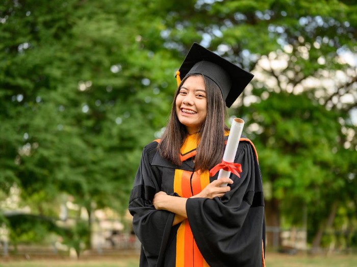 1151567114 A young female graduate with a scroll in her hands is smiling against the background of university graduates. Graduation.University gesture and people concept.