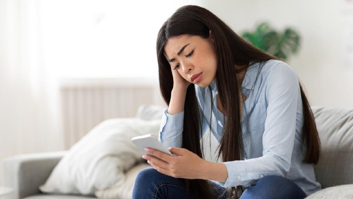 Waiting for his call. Upset asian girl looking sadly at mobile phone screen, sitting on sofa at home, copy space
