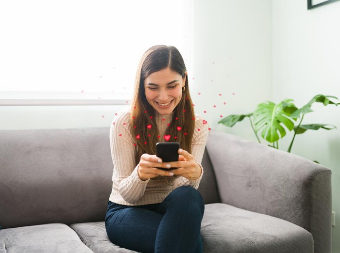 Young wife in love is sitting at home and texting her husband on her smartphone. Beautiful woman is sending loving messages to her partner