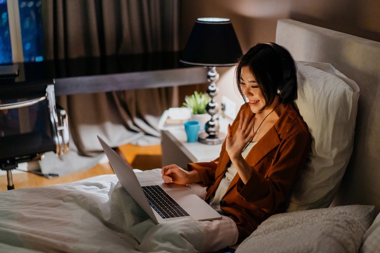 1296479137 Image of an Asian Chinese woman wearing headphone and video calling in bed with a laptop