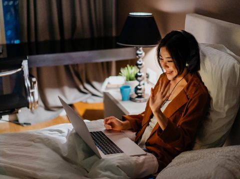 Image of an Asian Chinese woman wearing headphone and video calling in bed with a laptop