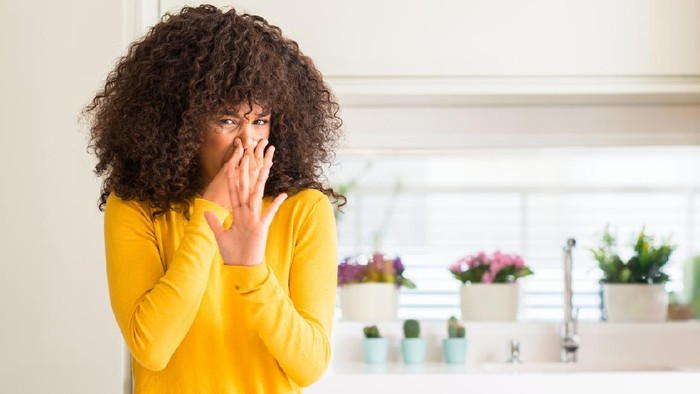 African american woman wearing yellow sweater at kitchen smelling something stinky and disgusting, intolerable smell, holding breath with fingers on nose. Bad smells concept.