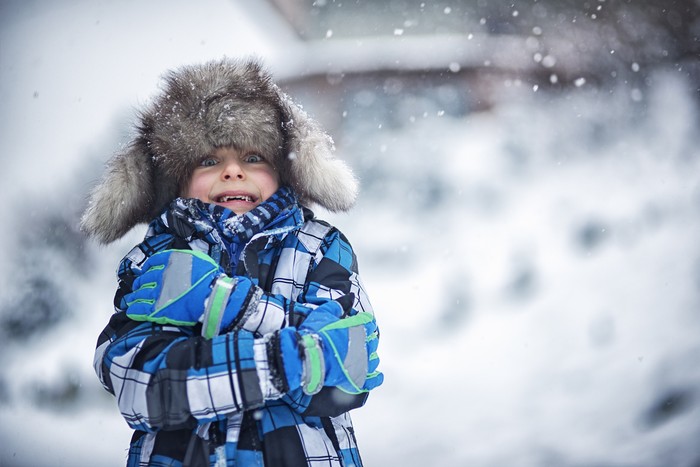 Portrait of a cute boy in big fake fur cap. Its snowing and freezing outside and the little boy is haking the winter day. Snow flakes are falling on the cap.
