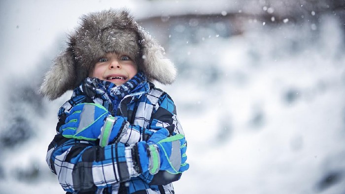 Portrait of a cute boy in big fake fur cap. Its snowing and freezing outside and the little boy is haking the winter day. Snow flakes are falling on the cap.