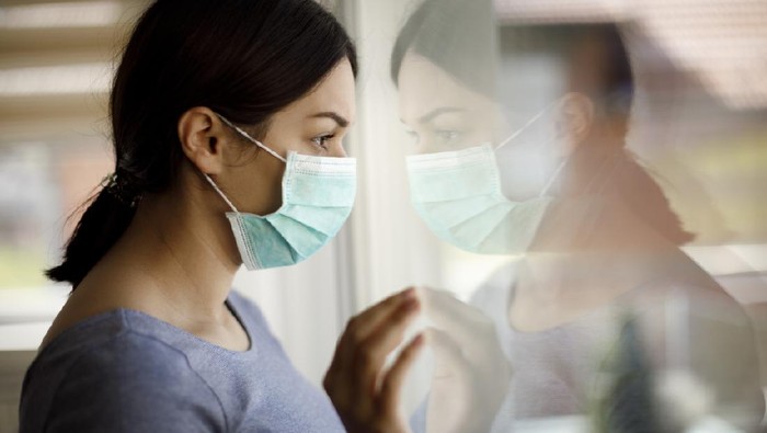 Portrait of sad young woman with face protective mask looking through the window at home