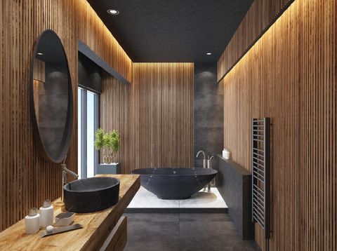 contemporary minimalist bathroom with wooden walls. oval mirror with black frame is on the wall with wooden battens.round stone washbasin and inox basin tap is on top of the long wooden vanity unit. freestanding black stone bathtub and floor mounted inox tapis on a platform with large white marble tiles. grey wall shelf and grey towel radiator on wooden wall. black ceiling with strip cove lighting with embedded spotlights.
