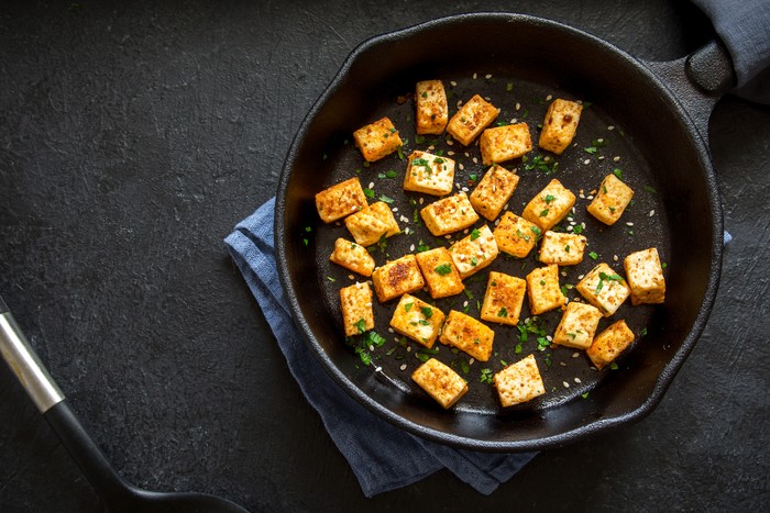 Fried tofu with sesame seeds and spices on cast iron pan, copy space. Healthy ingredient for cooking vegan vegetarian diet food. Roasted tofu over black background.