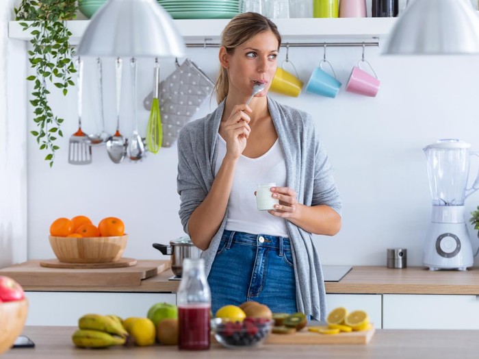 Ilustrasi wanita makan Shot of pretty young woman eating yogurt while standing in the kitchen at home.