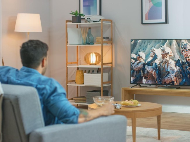 In the Living Room: Guy Relaxing on a Couch Watching War Movie on a TV. Modern Military Warfare Action with War Soldiers Shown on a Television.