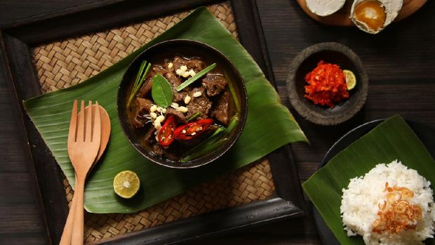 Rawon, the the popular and unique beef soup from East Java. Beef cubes cooked in rich broth flavored with native keluak fruit (pangium edule) and other herbs and spices. Topped with bean sprouts and chives. Accompanied with salted duck egg, red chili paste and steamed rice. The soup is served in a black ceramic bowl, then placed on a woven tray lined with banana leaf. A pair or wooden cutlery and lime half is also placed on the the tray. The accompanying side dishes and condiment are placed near the tray. The meal is arranged on a dark-colored wooden table.