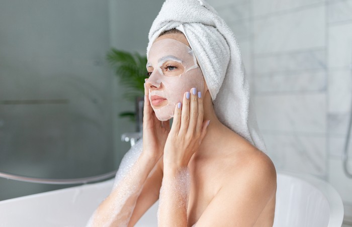 Ilustrasi Pemakaian Sheet Mask Beautiful young woman sitting in bathtub with sheet mask on face. Close up portrait of pretty girl in bath towel on head and doing beauty treatment. Home bathroom interior. Spa procedure. Skincare