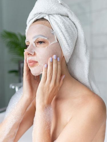 Beautiful young woman sitting in bathtub with sheet mask on face. Close up portrait of pretty girl in bath towel on head and doing beauty treatment. Home bathroom interior. Spa procedure. Skincare
