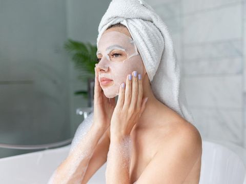 Beautiful young woman sitting in bathtub with sheet mask on face. Close up portrait of pretty girl in bath towel on head and doing beauty treatment. Home bathroom interior. Spa procedure. Skincare
