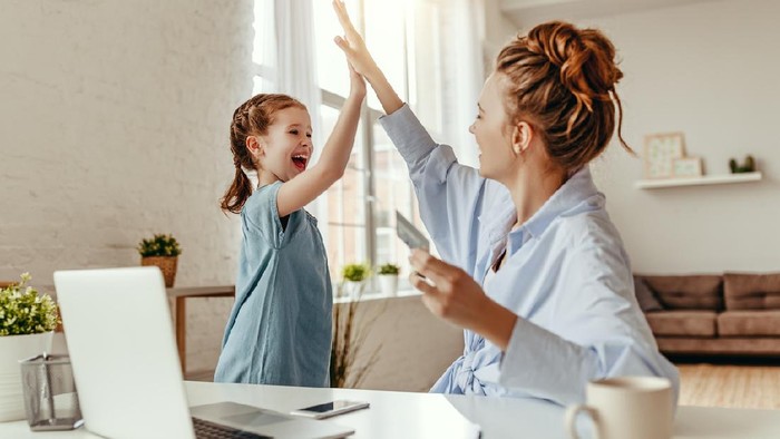 Low angle of excited small daughter giving high five to mother and screaming while celebrating successful online shopping using laptop at table in light living room