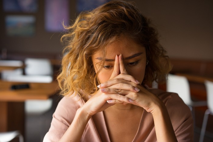 Closeup of sad young Asian woman at cafe leaning head on clasped hands and staring into vacancy. Tired freelancer feeling burnout. Stress and bad news concept