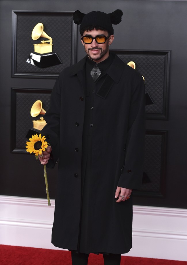Bad Bunny tampil dengan setelan serba hitam. Ia memadukan coat dan gaun lipit keluaran Burberry, ditambah boots hitam. Gayanya menjadi aneh karena topi bertelinga serta membawa setangkai bunga matahari. Foto: AP/Jordan Strauss
