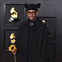 Bad Bunny tampil dengan setelan serba hitam. Ia memadukan coat dan gaun lipit keluaran Burberry, ditambah boots hitam. Gayanya menjadi aneh karena topi bertelinga serta membawa setangkai bunga matahari. Foto: AP/Jordan Strauss