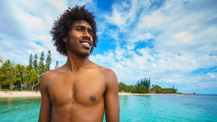 Happy smiling young pacific islander - kanak people, the original inhabitants of New Caledonia, looking over the turquoise lagoon at the beach of Noumea, New Caledonia. Real Native People Portrait. Noumea, New Caledonia, South West Pacific Ocean