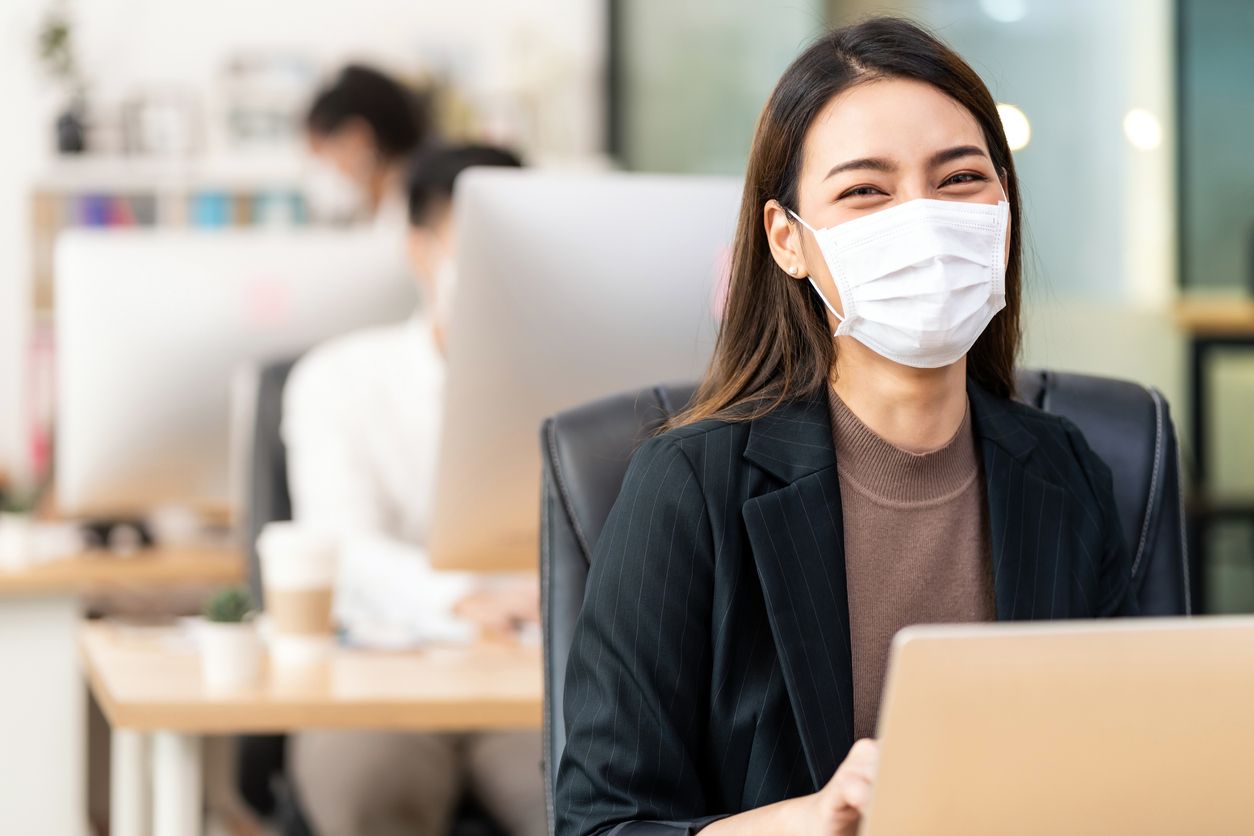 Portrait of asian office employee businesswoman wear protective face mask work in new normal office with interracial colleague in background as social distance practice prevent coronavirus COVID-19.