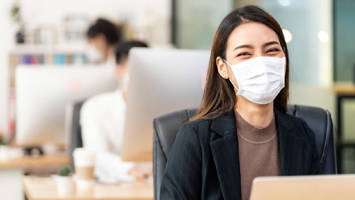 Portrait of asian office employee businesswoman wear protective face mask work in new normal office with interracial colleague in background as social distance practice prevent coronavirus COVID-19.