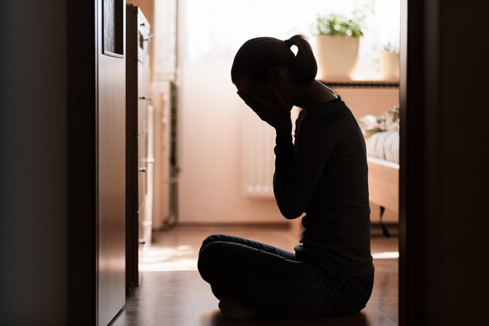 1147847234 Sad young woman sitting on room floor crying with hand over face