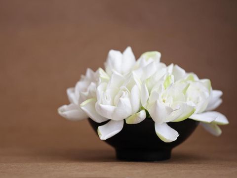 A DSLR still life photo of a bouquet of fresh white gardenia flowers in a small black ceramic bowl. Background made of brown rustic paper. Selective focus on the flower of the foreground. Soft natural light. Copy space.