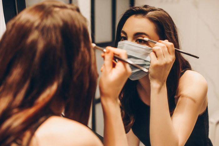 1253160478 selective focus of young woman in medical mask and black dress applying eye shadow in bathroom