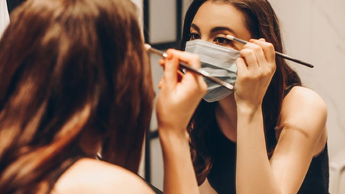 selective focus of young woman in medical mask and black dress applying eye shadow in bathroom