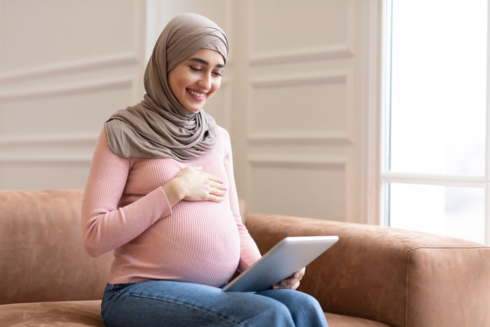Joyful Pregnant Muslim Lady Using Digital Tablet Browsing Internet Sitting On Couch At Home. Pregnancy Lifestyle Concept. Arab Mother-To-Be Using Computer. Copy Space For Text