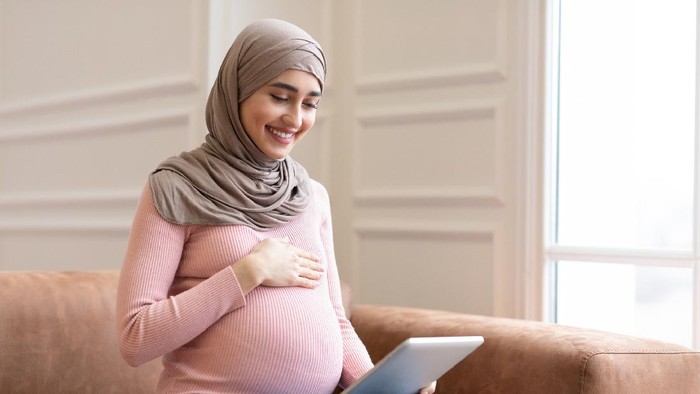 Joyful Pregnant Muslim Lady Using Digital Tablet Browsing Internet Sitting On Couch At Home. Pregnancy Lifestyle Concept. Arab Mother-To-Be Using Computer. Copy Space For Text