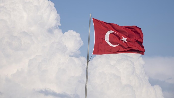 Turkish Flag on cloudy blue sky.The flag is waving due to heavy wind.On the background white clouds are seen.Up right side of frame is blue sky.No people are seen in frame.