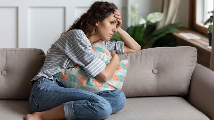 1207723226 Frustrated young lady sitting on sofa, cuddling pillow, looking away at window. Lost in thoughts unhappy stressed millennial woman regretting of wrong decision, spending time alone in living room.