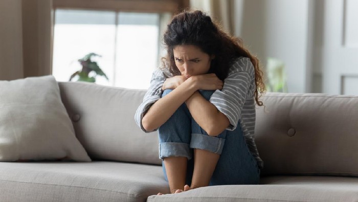 1207723232 Stressed young woman embracing knees, sitting alone on couch in living room, feeling depressed. Unhappy desperate millennial curly lady regretting of decision, thinking of personal problems at home.