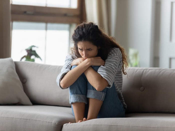 1207723232 Stressed young woman embracing knees, sitting alone on couch in living room, feeling depressed. Unhappy desperate millennial curly lady regretting of decision, thinking of personal problems at home.