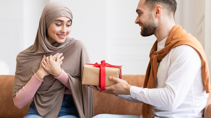 Valentines Day. Arab Man Surprising Wife With A Gift Giving Wrapped Present Box To Her Sitting On Couch At Home. Birthday Holiday Celebration, Romantic Presents And Gifts. Panorama
