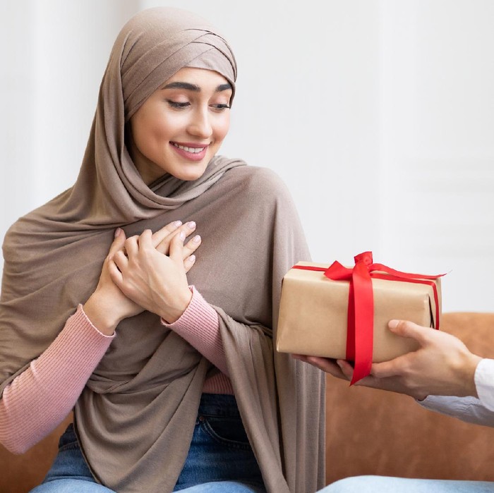 Valentines Day. Arab Man Surprising Wife With A Gift Giving Wrapped Present Box To Her Sitting On Couch At Home. Birthday Holiday Celebration, Romantic Presents And Gifts. Panorama