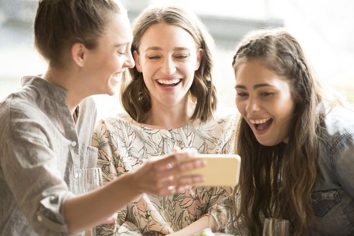 504880396 Three women looking at smartphone, smiling and laughing. Three female friends using social media, having fun, togetherness