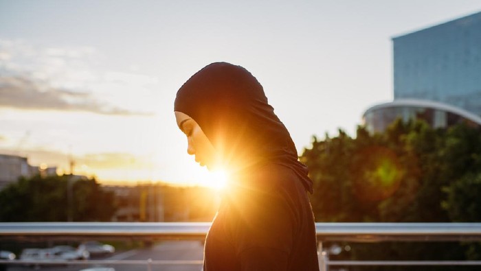 Arabic woman runner outdoors in the morning with bright sunlight. Side view of young muslim woman wearing hijab resting after workout outdoors in the city.