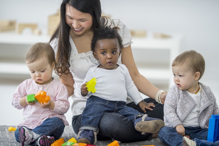 Pengasuh Anak Crazy Rich A group of babies are sitting with their babysitter on the carpet. The babies are playing with colorful toy blocks.