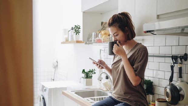 Teenage asian woman drinking coffee while sitting on kitchen counter and working on smart phone in morning at home.