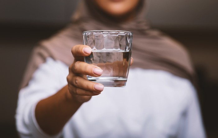 Unrecognizable Muslim Woman Offering Glass Of Water To Camera Standing In Modern Kitchen At Home, Wearing Hijab. Healthy Hydration, Drink More Water And Stay Hydrated. Selective Focus, Cropped