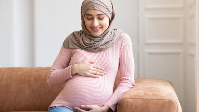 Cheerful Pregnant Muslim Lady In Hijab Posing Embracing Belly Sitting On Couch At Home. Young Arab Moms Portrait, Awaiting Baby. Pregnancy Happiness And Childbirth Concept