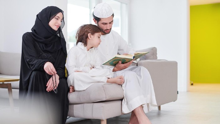 Traditional muslim family parents with children reading Quran and praying together on the sofa before iftar dinner during a ramadan feast at home